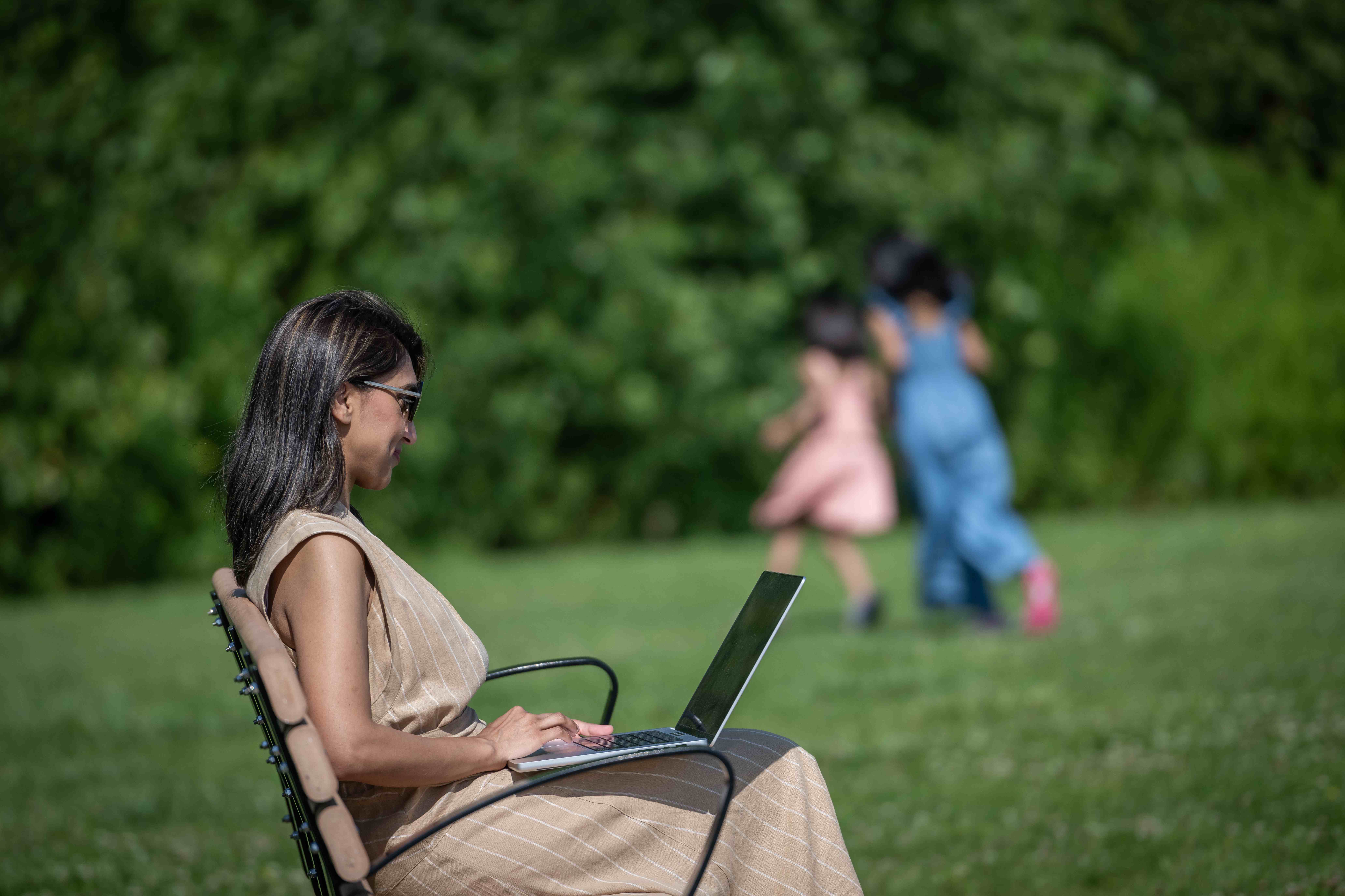 student sits outside on laptop while children play in background