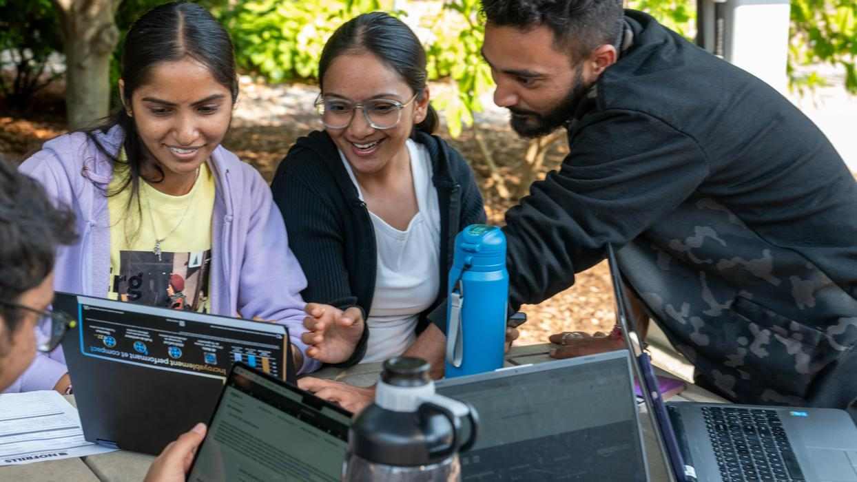 students sit around an outdoor table, pointing at laptop